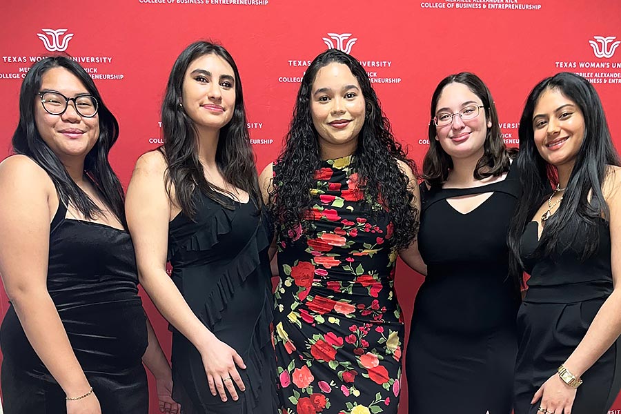 five business students, dressed in formal wear, stand in front of a maroon backdrop