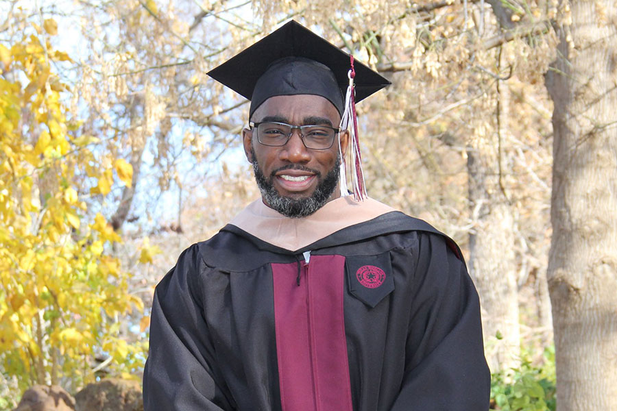 graduate in cap and gown stands outside in front of trees and foliage
