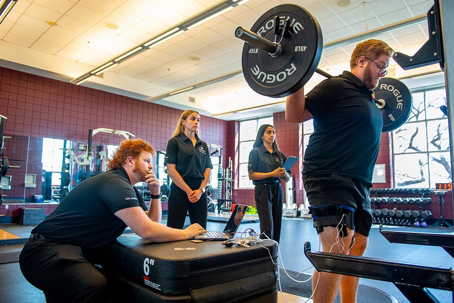 a student in black shirt holds a bar bell with weights on it with three students watching him in a guy