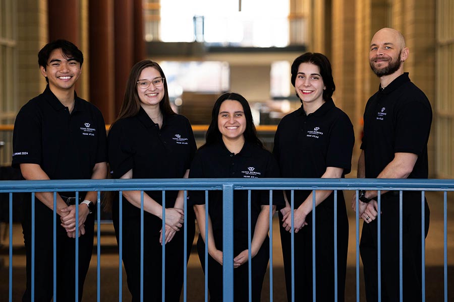 Five students, dressed in black, stand next to one another behind an indoor railing