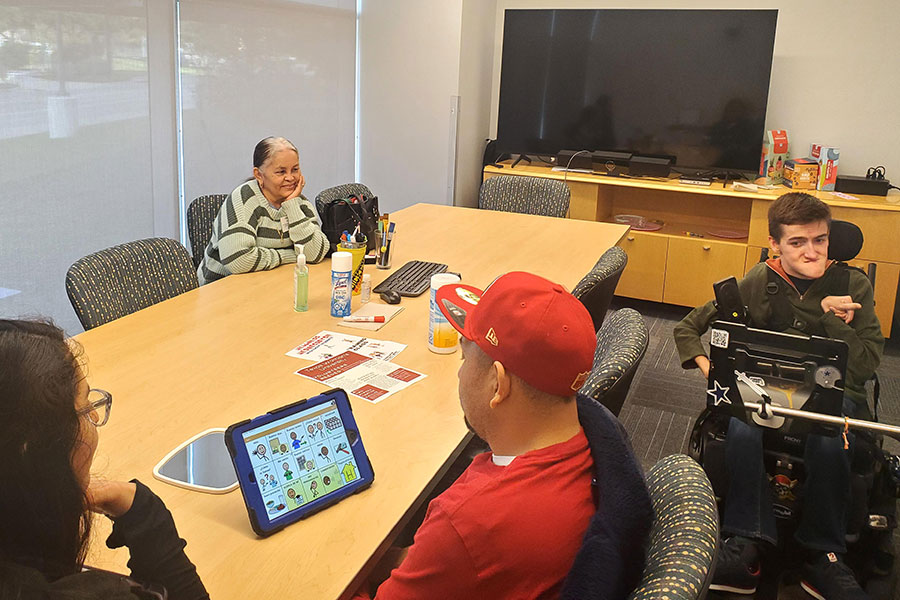 A speech-language pathology graduate student sits next to Stroke Center client who has tablet in front of him on table. Watson Polk sits next to table